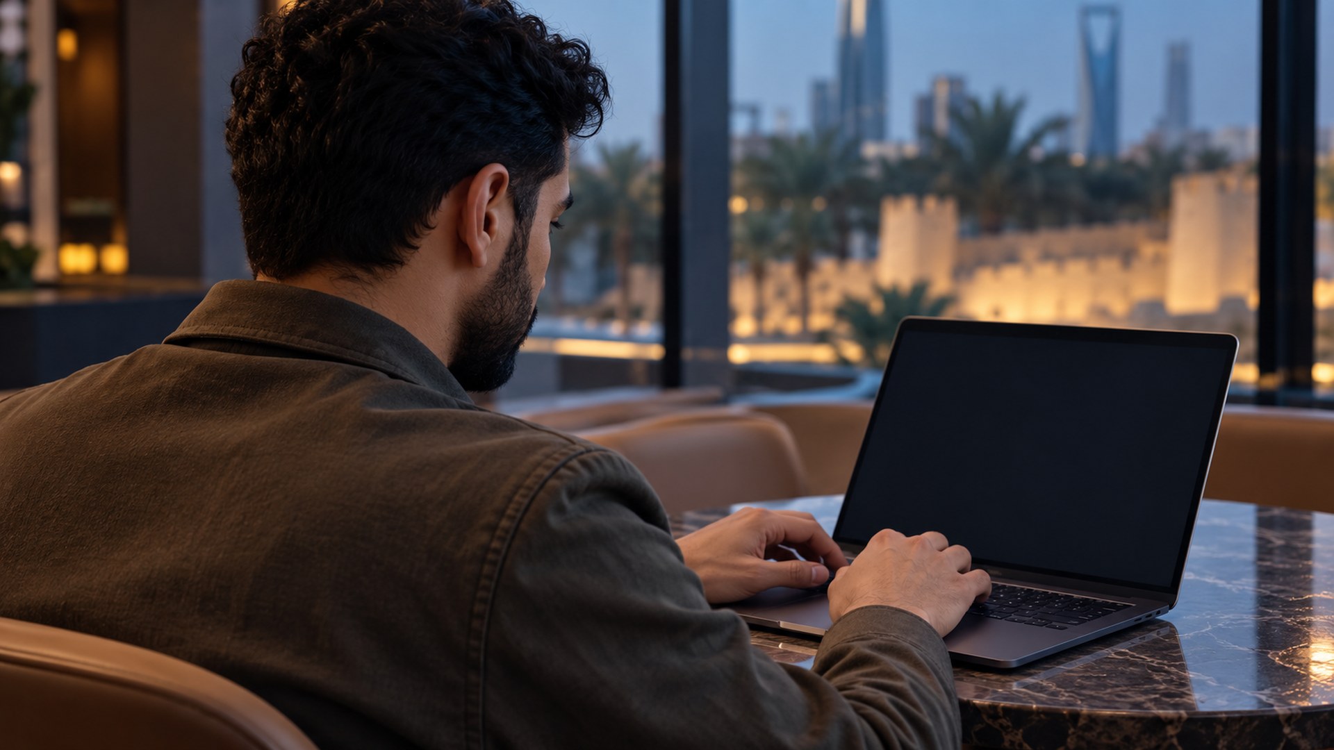 رجل سعودي يكتب عبارة بحث على لابتوبه في مقهى هادئ، من فوق كتفه