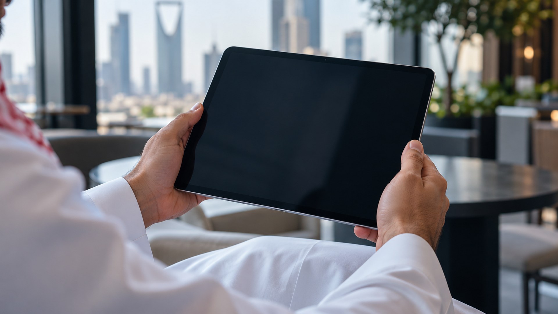 يدا رجل سعودي تمسكان جهازاً لوحياً في مقهى مكتبي حديث بضوء النهار
