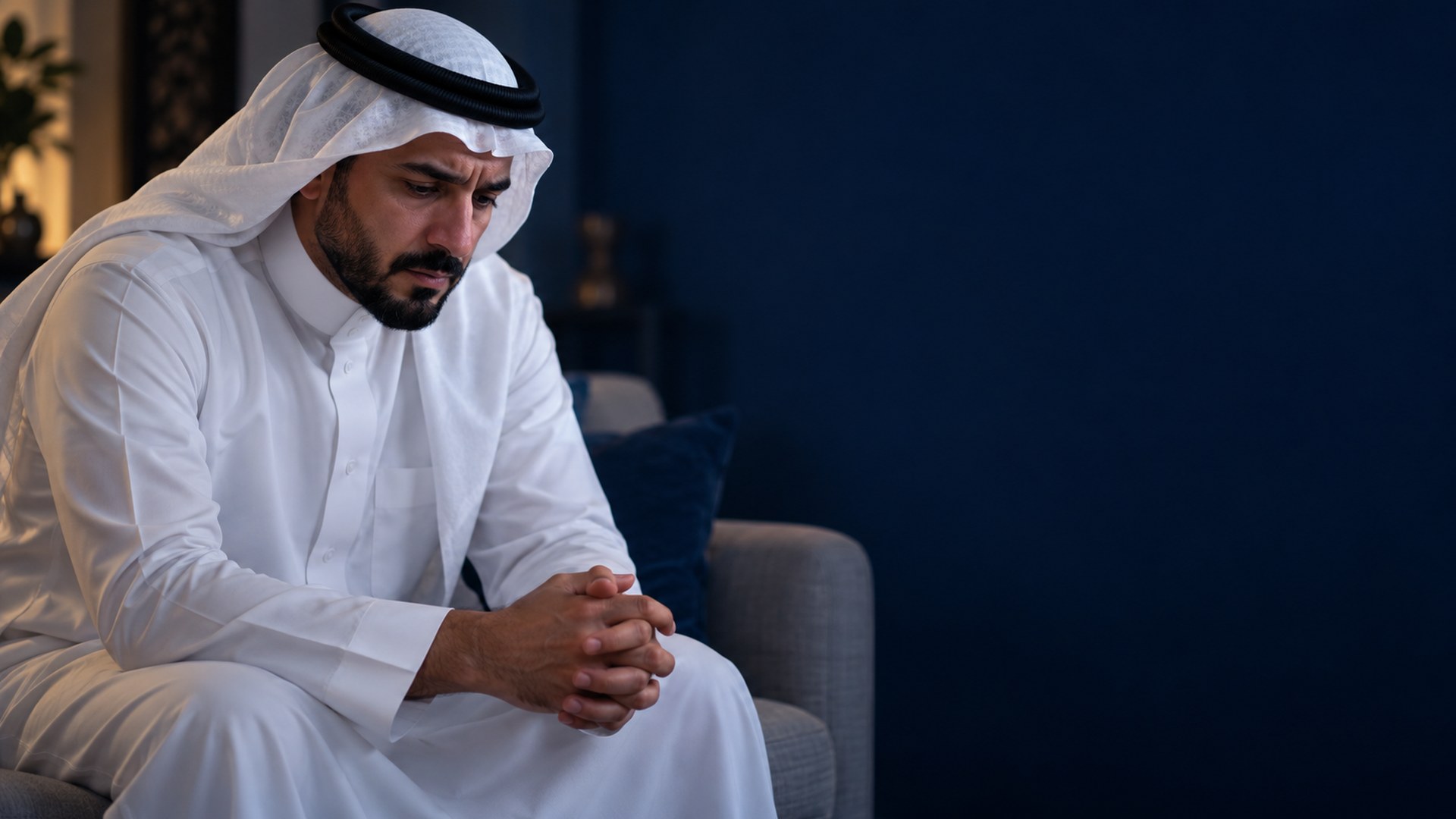 رجل سعودي على طرف الكنب بنظرة حزينة ويدين مشبوكتين في بيت هادئ