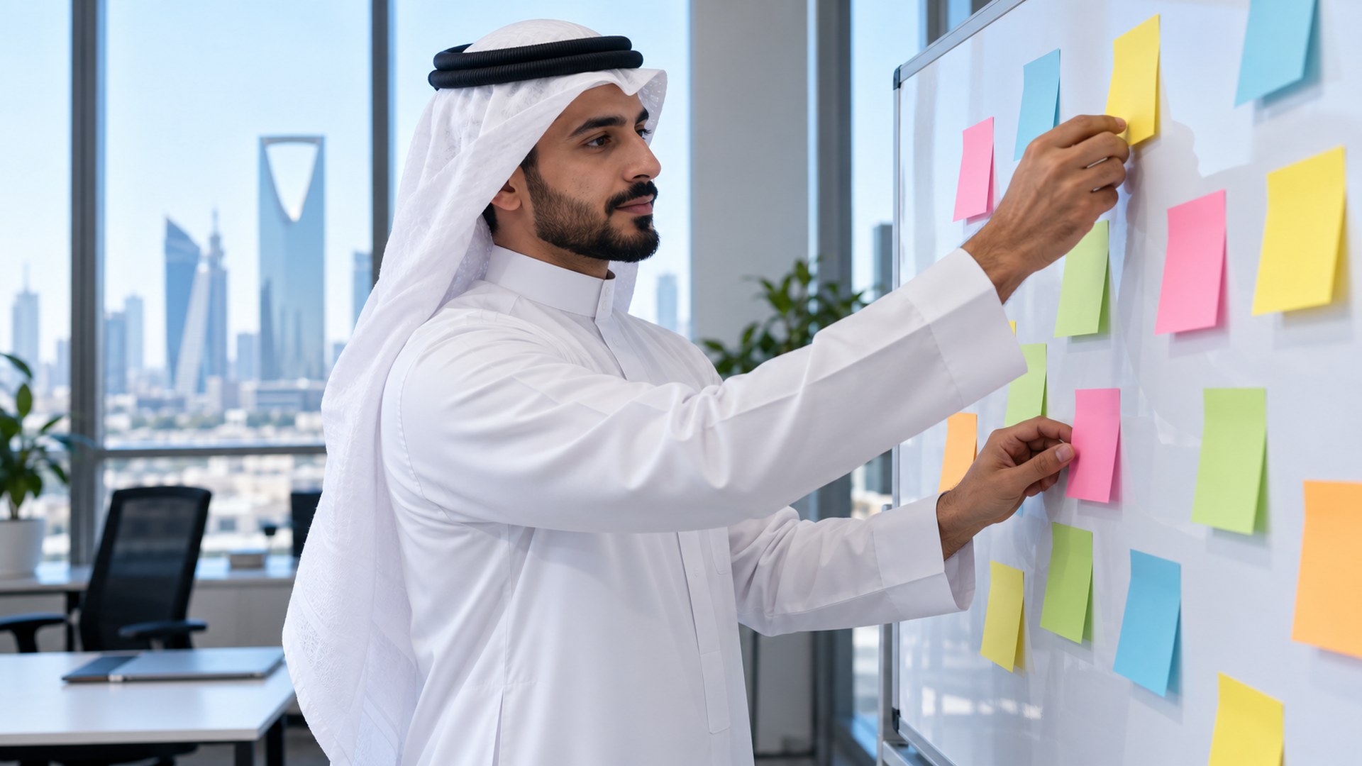 رجل سعودي بالثوب يرتّب أوراقاً لاصقة ملونة على سبورة في مكتب مضيء