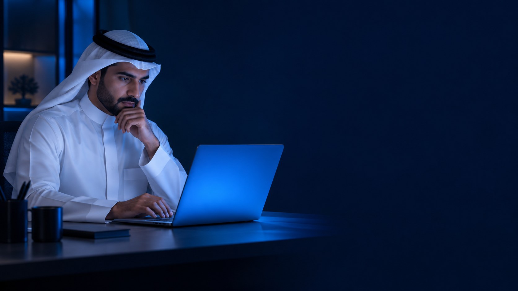 رجل سعودي بالثوب والغترة يتأمل شاشة لابتوبه في مكتب حديث بإضاءة زرقاء هادئة