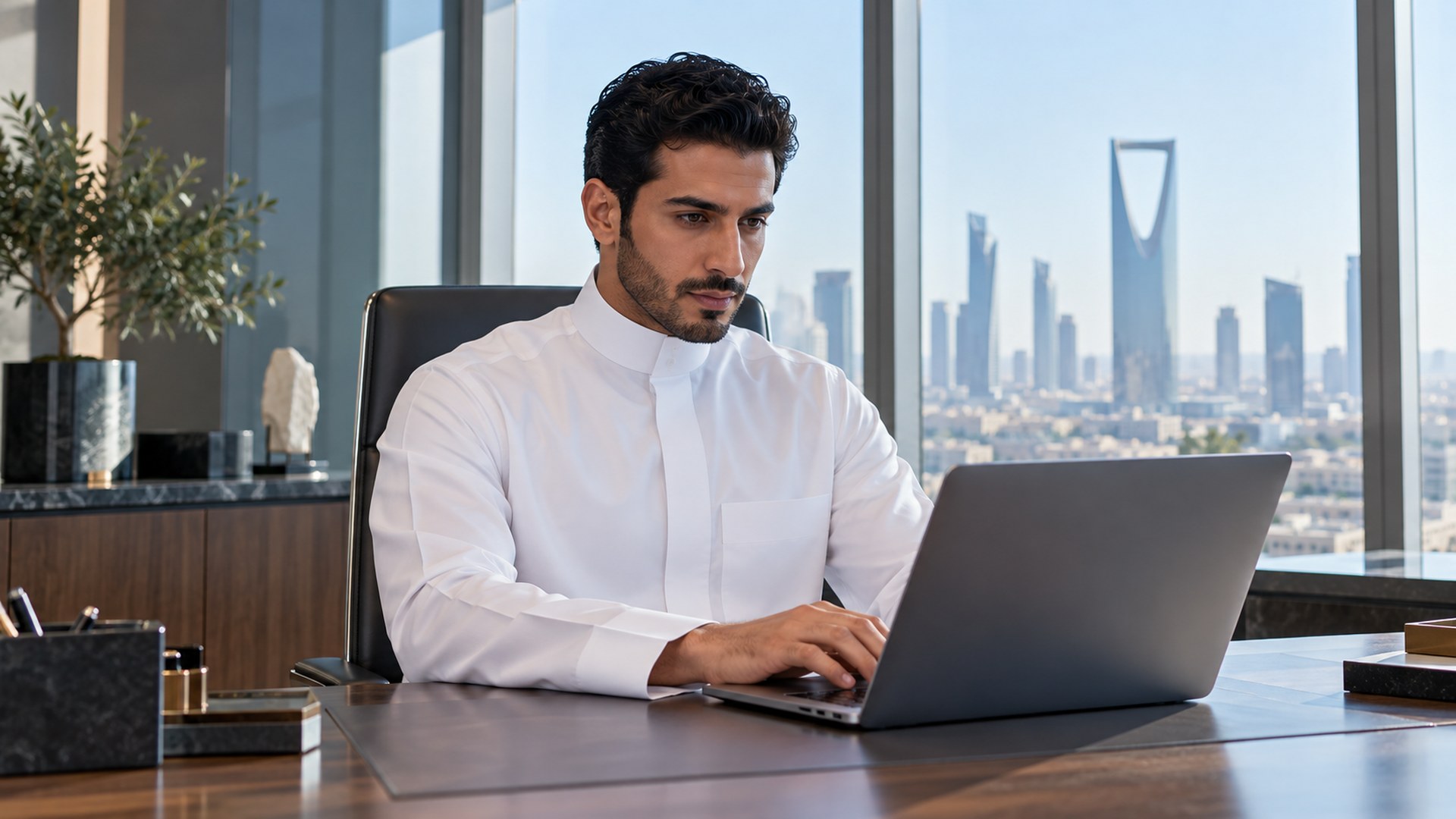 موظف سعودي بثوب أبيض يكتب طلباً على لابتوب أنيق في مكتب مضيء