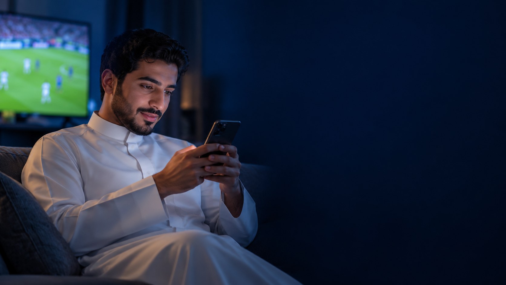 شاب سعودي على الكنب ليلاً ينظر لجواله بفضول ومباراة على التلفزيون خلفه