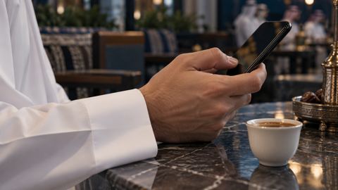 يد تتصفح الجوال على طاولة مقهى بجانب فنجان قهوة عربية صغير