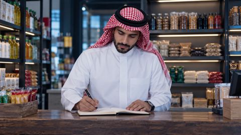 صاحب محل سعودي يكتب قائمة في دفتر عند الطاولة وخلفه رفوف مرتبة