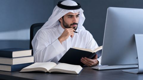 محرر سعودي يراجع مسودة مقال على شاشة الكمبيوتر وبجانبه كتب مراجع