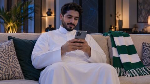 مشجع سعودي يتفقد جواله بجانب وشاح كرة قدم على الكنب