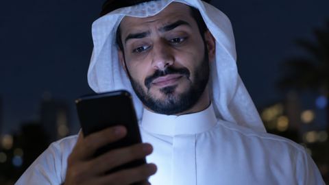 وجه رجل سعودي يضيئه جواله ليلاً وهو يرفع حاجبيه شاكاً