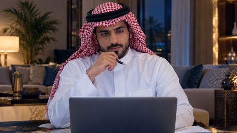 رجل سعودي بالشماغ الأحمر يتوقف متأملاً أمام اللابتوب والقلم في يده