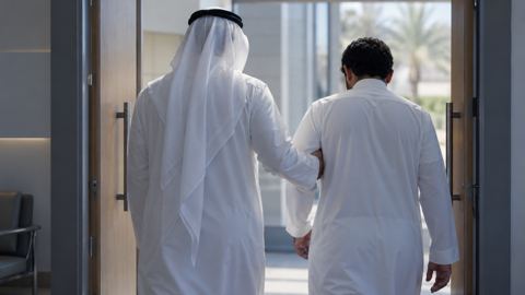 رجل سعودي يسند صديقه وهما يخرجان من باب العيادة، من الخلف