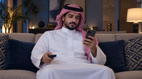 رجل سعودي على الكنب يتابع جدول البرامج على جواله والريموت في يده