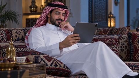 رجل سعودي متكئ في المجلس يقرأ دليلاً على جهازه اللوحي باهتمام