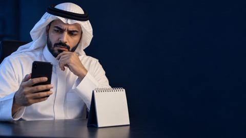 رجل سعودي بثوب أبيض ينظر لجواله متسائلاً بجانب تقويم مكتبي