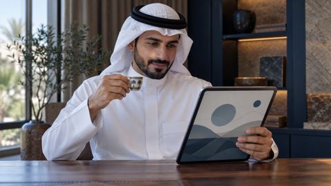رجل سعودي يقرأ دليلاً على جهاز لوحي ويمسك فنجان قهوة عربية