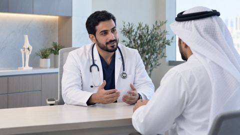 طبيب عظام سعودي بمعطف أبيض يناقش خيارات العلاج مع مريض في عيادة مضيئة