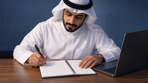 شخص سعودي يكتب قائمة واضحة في دفتر بجانب لابتوب على مكتب خشبي