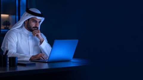 رجل سعودي بالثوب والغترة يتأمل شاشة لابتوبه في مكتب حديث بإضاءة زرقاء هادئة