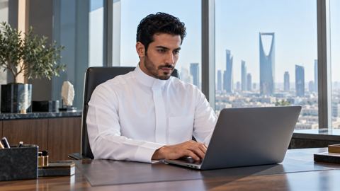 موظف سعودي بثوب أبيض يكتب طلباً على لابتوب أنيق في مكتب مضيء