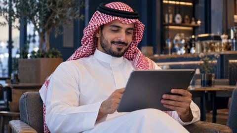 قارئ سعودي يبتسم وهو يقرأ مقالاً مفيداً على جهاز لوحي في مقهى