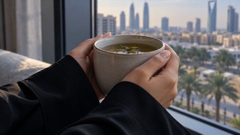 يدا امرأة بأكمام طويلة تمسكان كوب شاي أعشاب بجانب النافذة