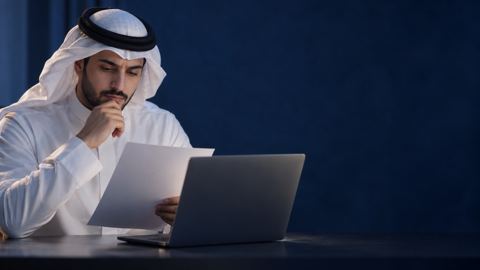 شاب سعودي بالثوب يدرس ورقة أكاديمية بجانب لابتوبه بتأمل