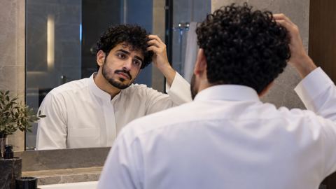 شاب سعودي بثوب أبيض يتفقد شعره المجعد الجاف في مرآة الحمام