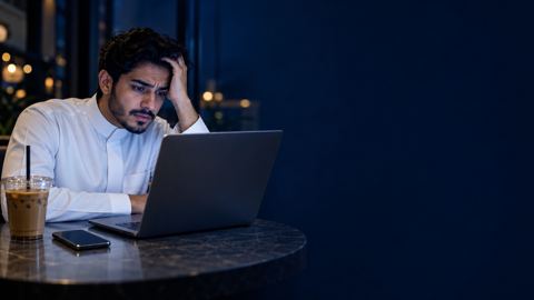 شاب سعودي متعب ومحتار أمام لابتوبه في مقهى ليلاً وبجانبه قهوة باردة