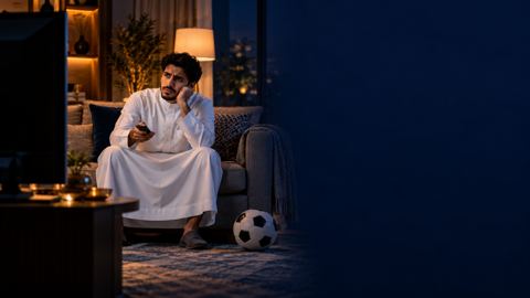 شاب سعودي على الكنب يمسك الريموت محتاراً أمام تلفزيون مطفأ وبجانبه كرة